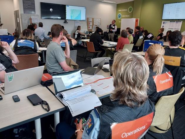 Group of individuals in a meeting room with laptops and documents open, attending a presentation about emergency operations. Visible on their vests are 'Operations' labels.